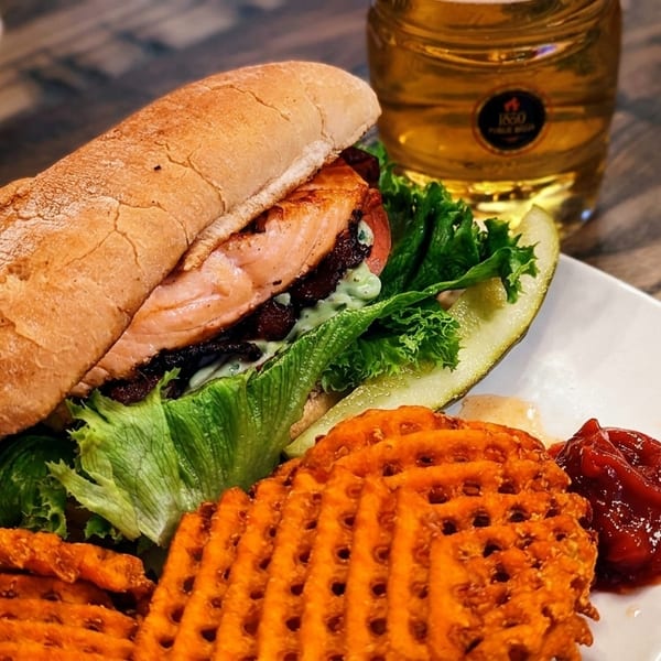 Salmon sandwich with lettuce, tomato, bacon, waffle fries, pickle, ketchup, and a beer.