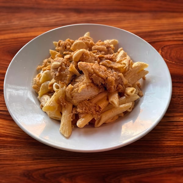 A white plate filled with penne pasta, creamy sauce, and crispy fried chicken pieces on a wooden table.