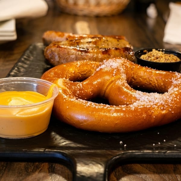A salted pretzel, two sausages, cheese dip, and grainy mustard on a black serving board.