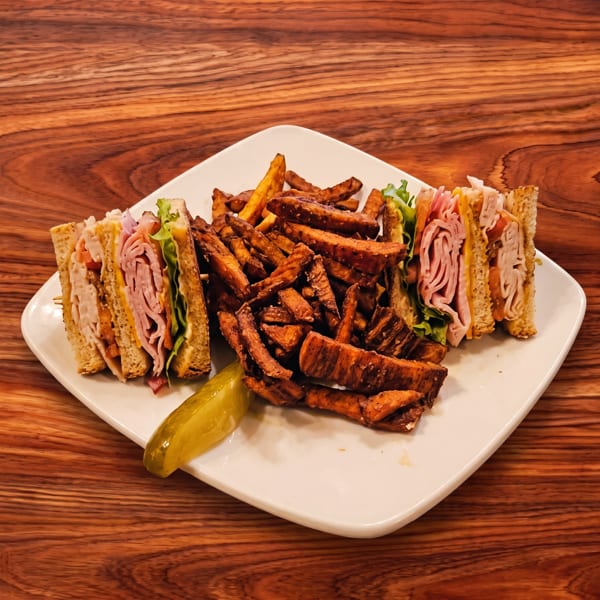 Club sandwich halves with sweet potato fries and a pickle spear on a white plate.