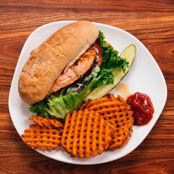 Grilled salmon sandwich on a white plate with waffle fries, ketchup, and a pickle spear.