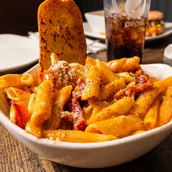 Penne pasta with meatballs, bacon, and garlic bread, with a Pepsi in the background.